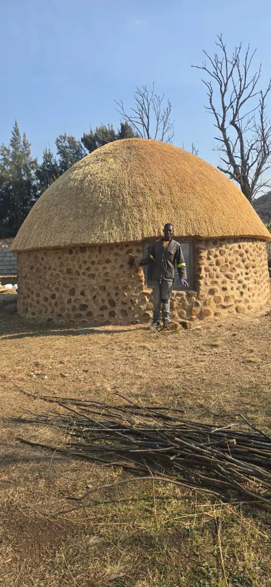A man standing in front of a round stone house with a large thatched roof